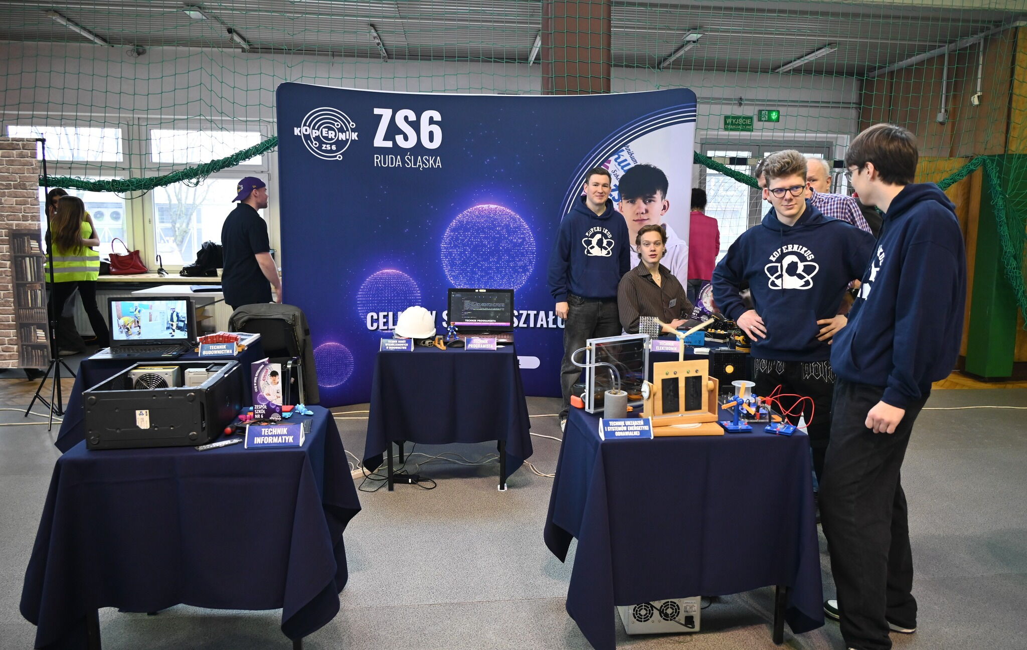 w pomieszczeniu stoisko szkolne, młodzież w bluzach z logo szkoły prezentuje urządzenia