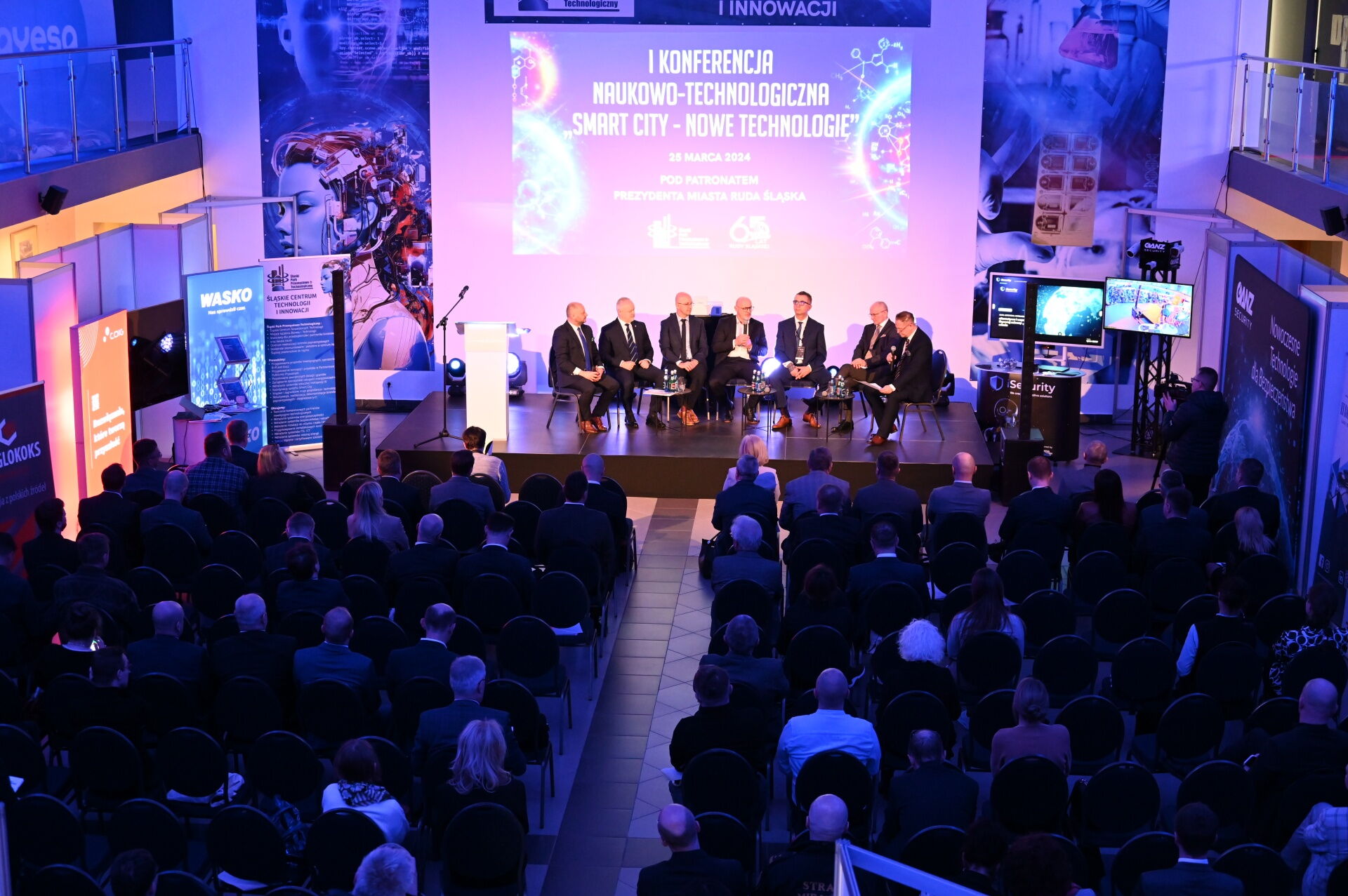 Widok z góry na salę konferencyjną: widownia, prelegenci na scenie, ekran z tekstem "I Konferencja Naukowo-Technologiczna „Smart City – nowe technologie”".