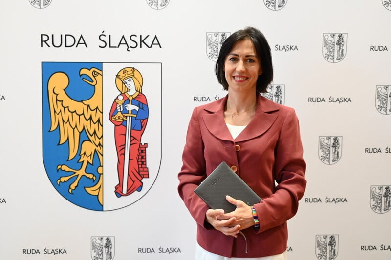 Młoda kobieta w eleganckim ubraniu trzyma w ręce kalendarz książkowy. Za nią ścianka, na której są napisy Ruda Śląska oraz zamieszczony jest herb miasta (z lewej strony połowa orła, z prawej postać św. Barbary).