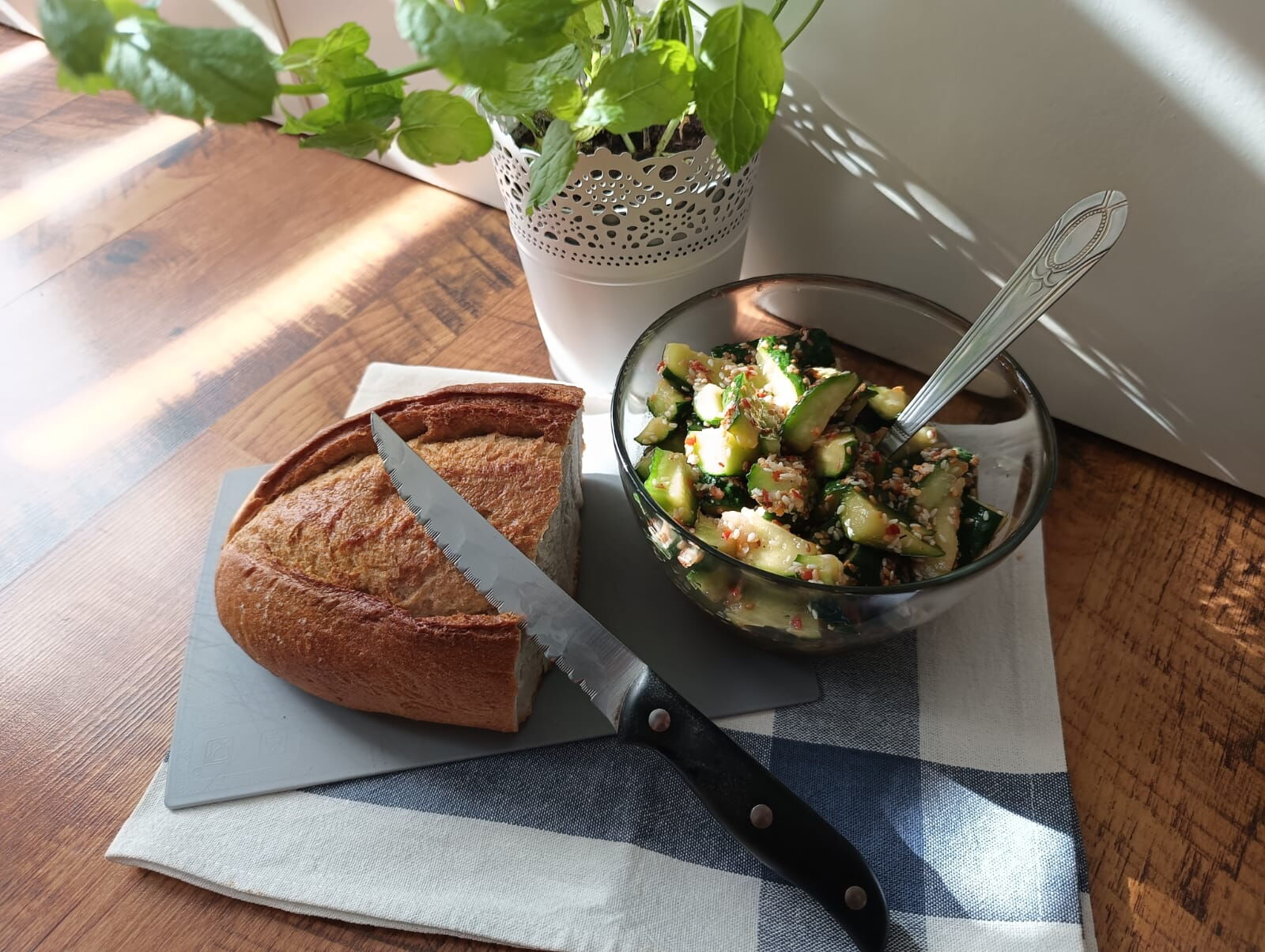 Na blacie leży ścierka, na niej pół chleba, miseczka z przyprawionymi ogórkami, obok stoi mięta w doniczce.