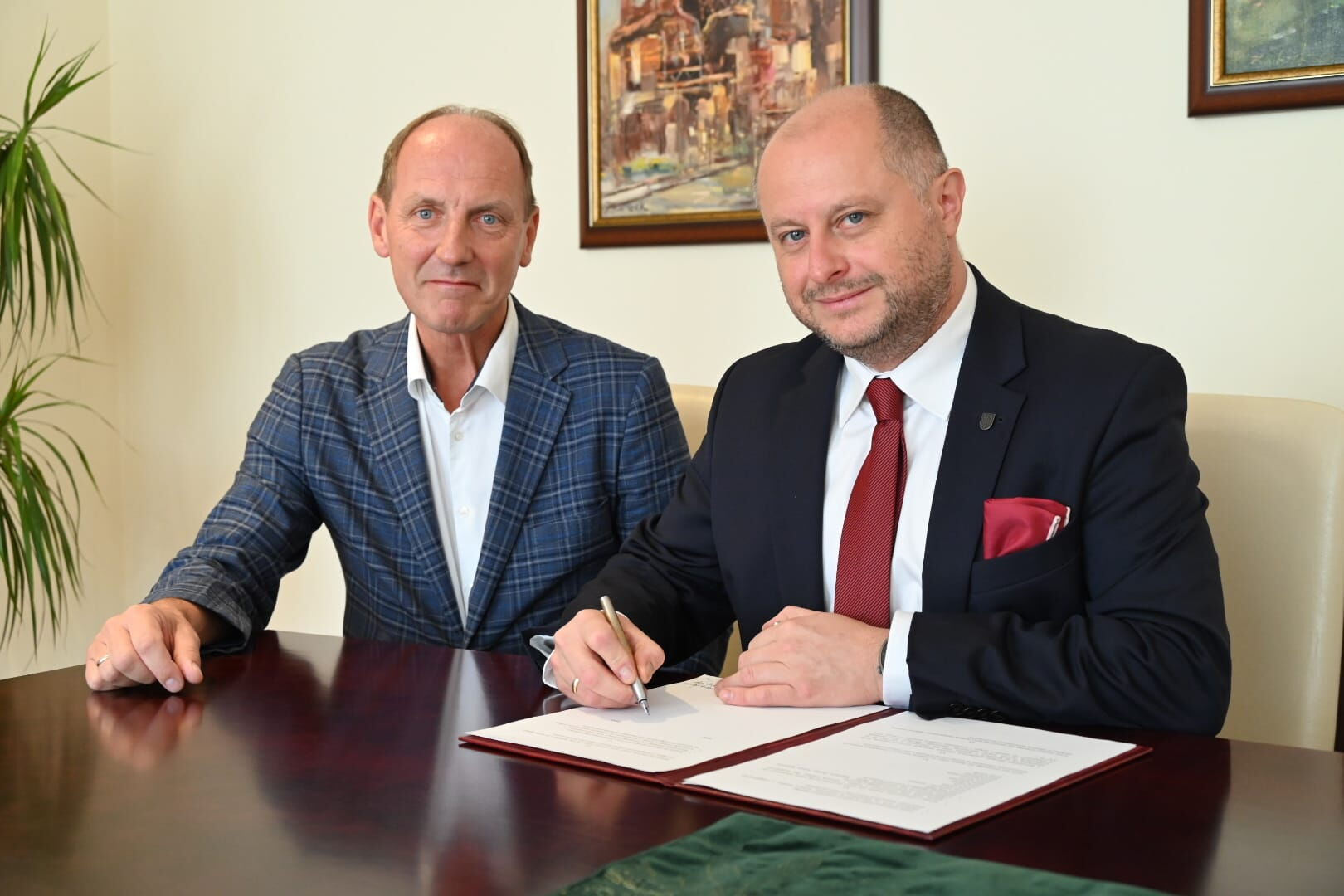 Dwóch mężczyzn w garniturach siedzi przy stole - jeden z nich podpisuje dokumenty. W tle na ścianie wiszą dwa obrazy.