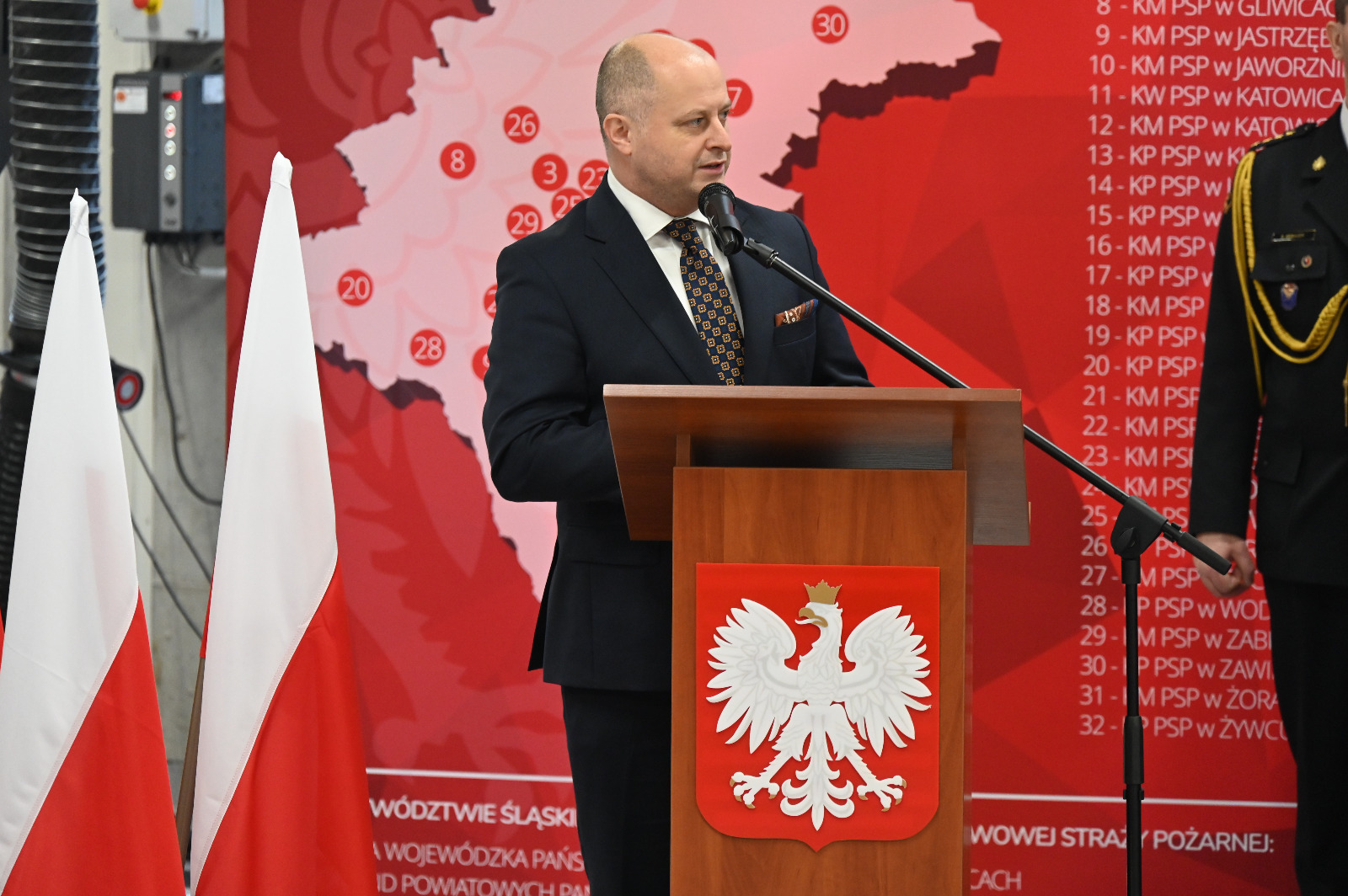 Mężczyzna w garniturze przy mównicy z godłem Polski. Obok flagi Polski. W tle ścianka w mapą rozmieszczenia komend Straży Pożarnej.