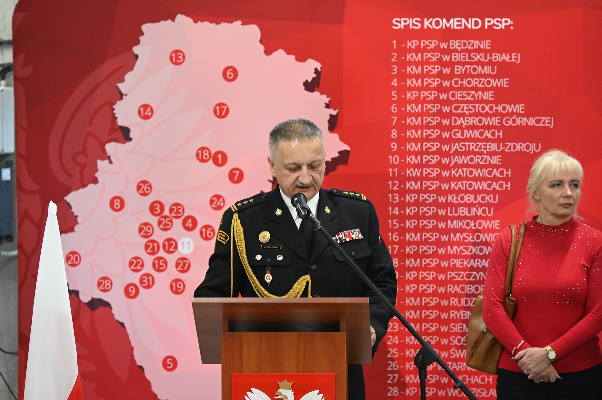 Mężczyzna w mundurze strażackim przy mównicy z godłem Polski. Obok flagi Polski. W tle ścianka w mapą rozmieszczenia komend Straży Pożarnej oraz kobieta w czerwonym swetrze w mundurze strażackim.