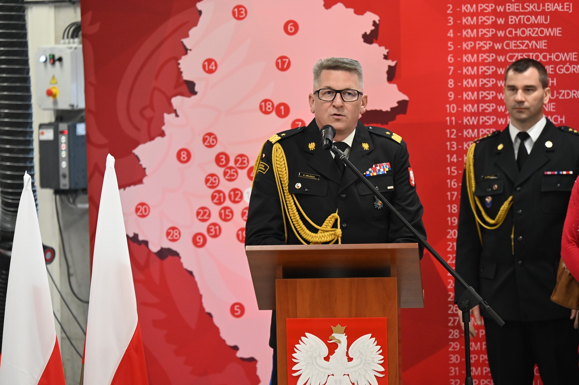 Mężczyzna w mundurze strażackim przy mównicy z godłem Polski. Obok flagi Polski. W tle ścianka w mapą rozmieszczenia komend Straży Pożarnej oraz mężczyzna w mundurze strażackim.
