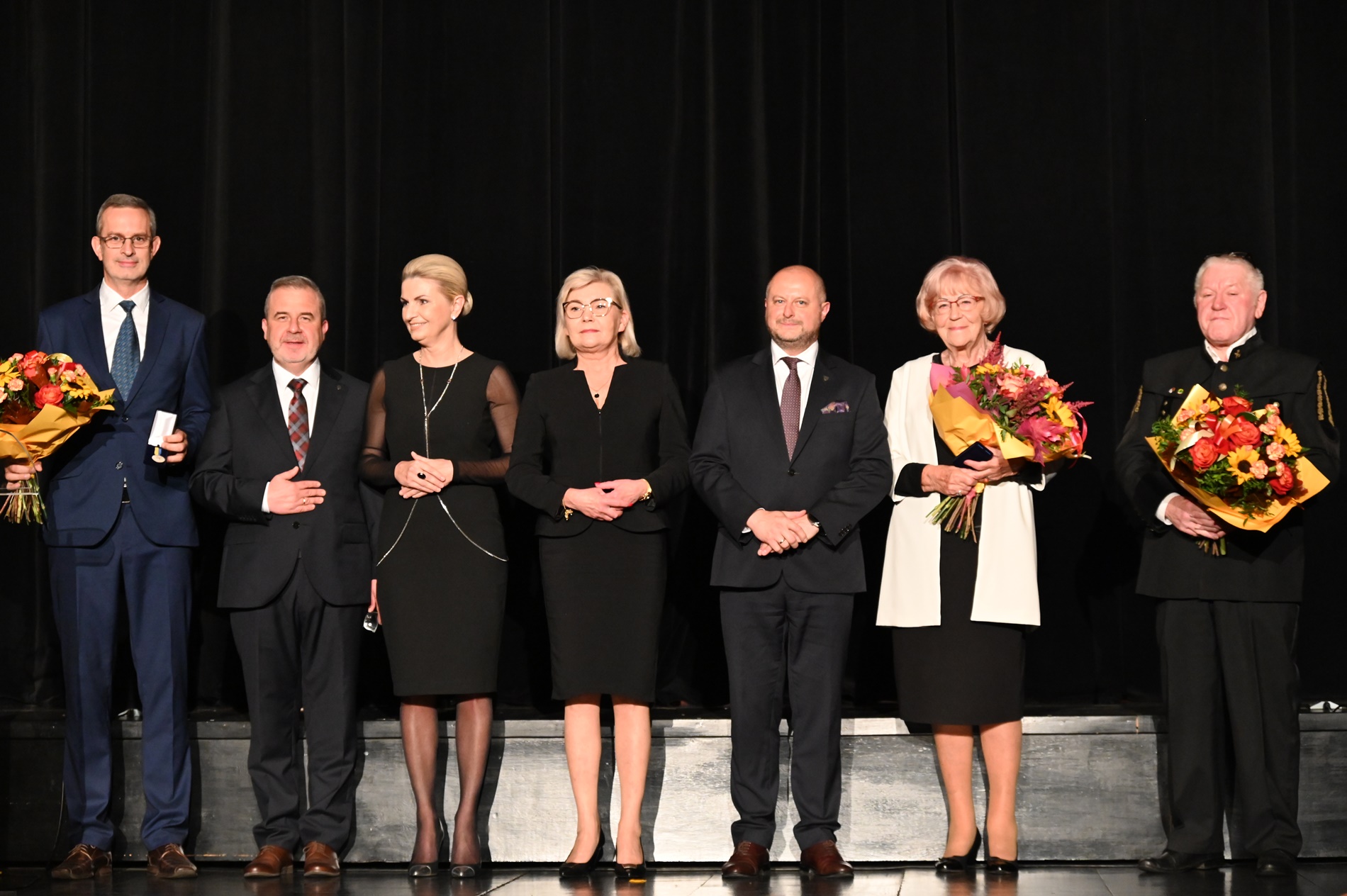 Grupa kilku osób w eleganckich strojach stoi na scenie. Trzy osoby trzymają bukiety kwiatów.