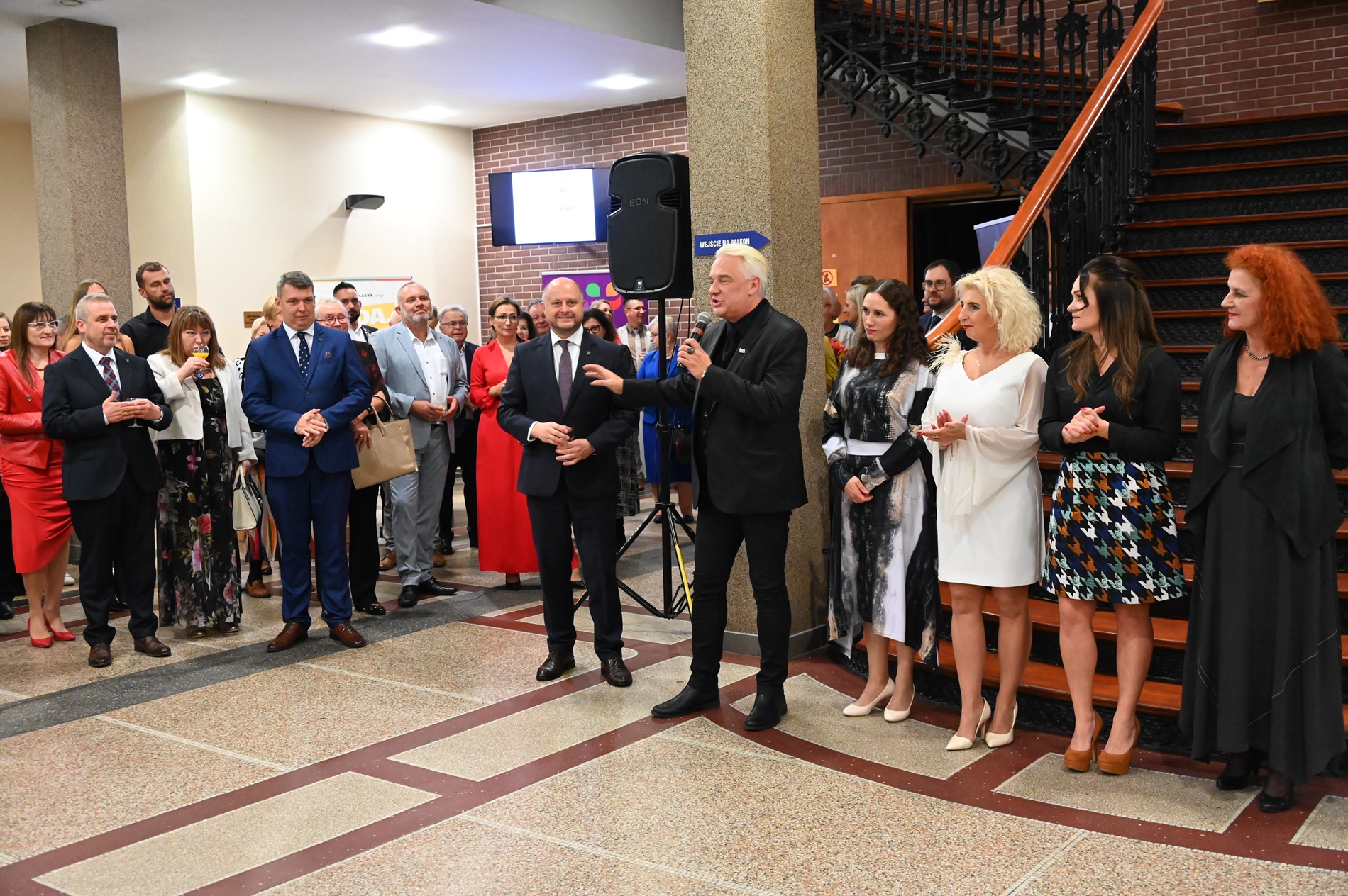 Sala - duża grupa osób stoi, mężczyzna mówi do mikrofonu. W tle schody prowadzące do góry.