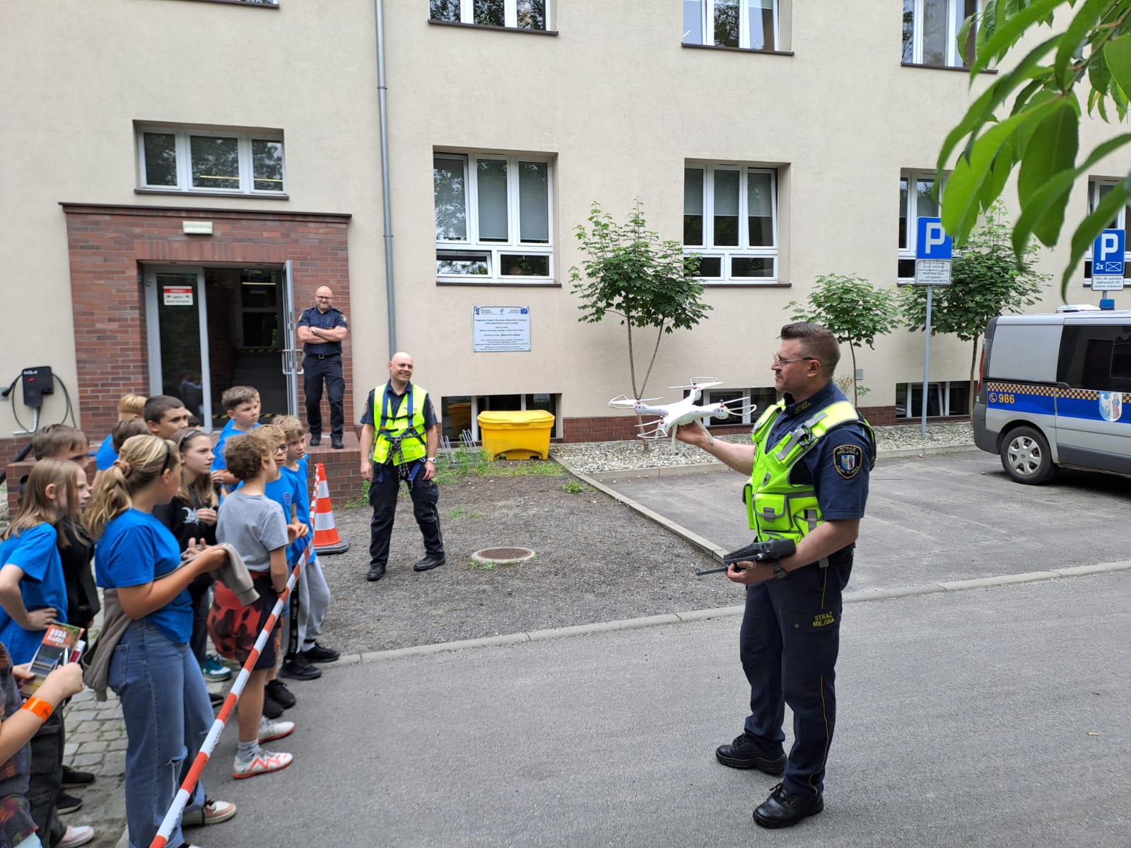 Strażnik miejski prezentuje drona grupie dzieci. W tle dwóch strażników miejskich, samochód straży miejskiej i budynek.