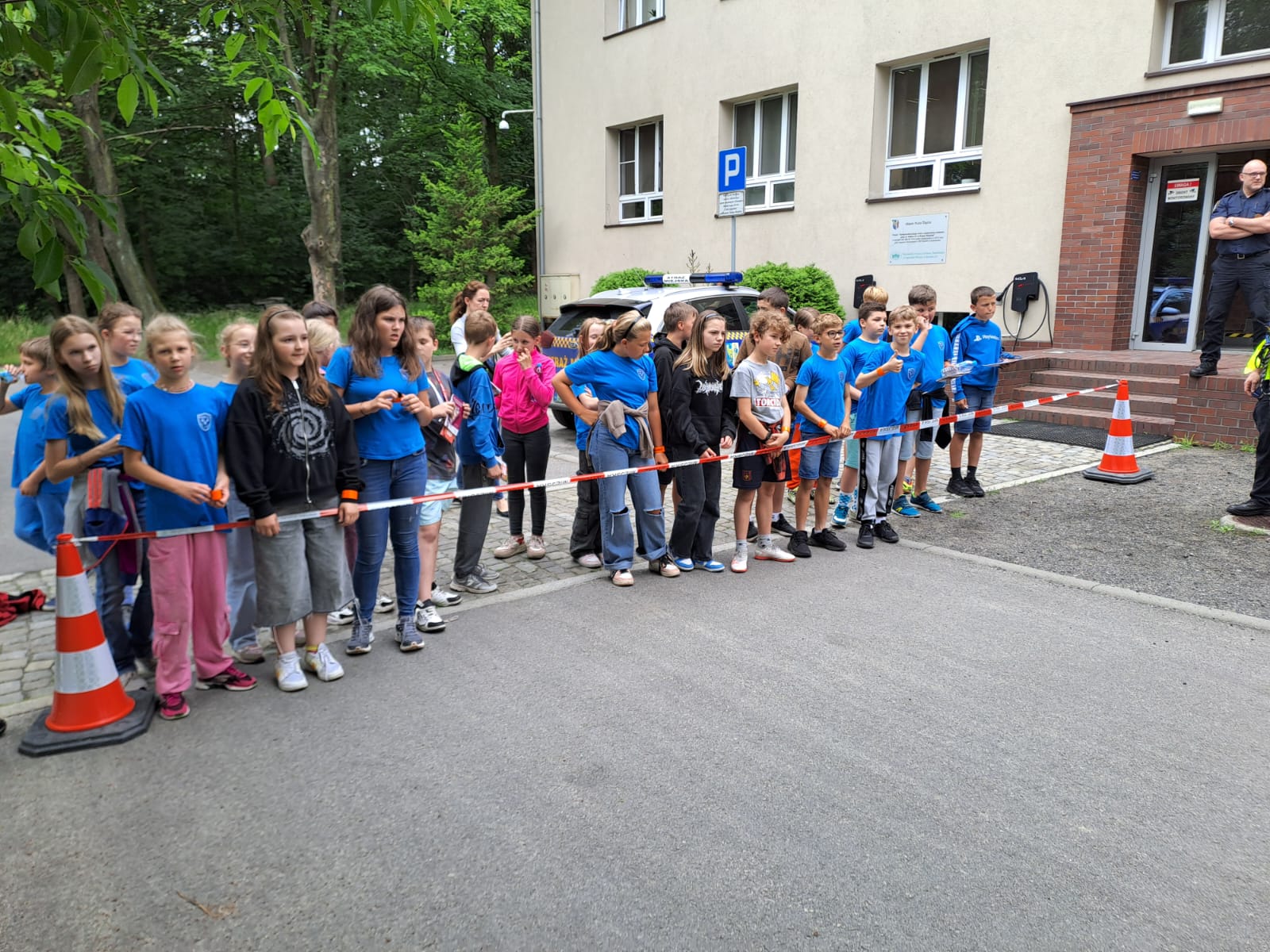 Grupa dzieci przed taśmą rozciągniętą między pachołkami. W tle strażnik miejski i budynek.